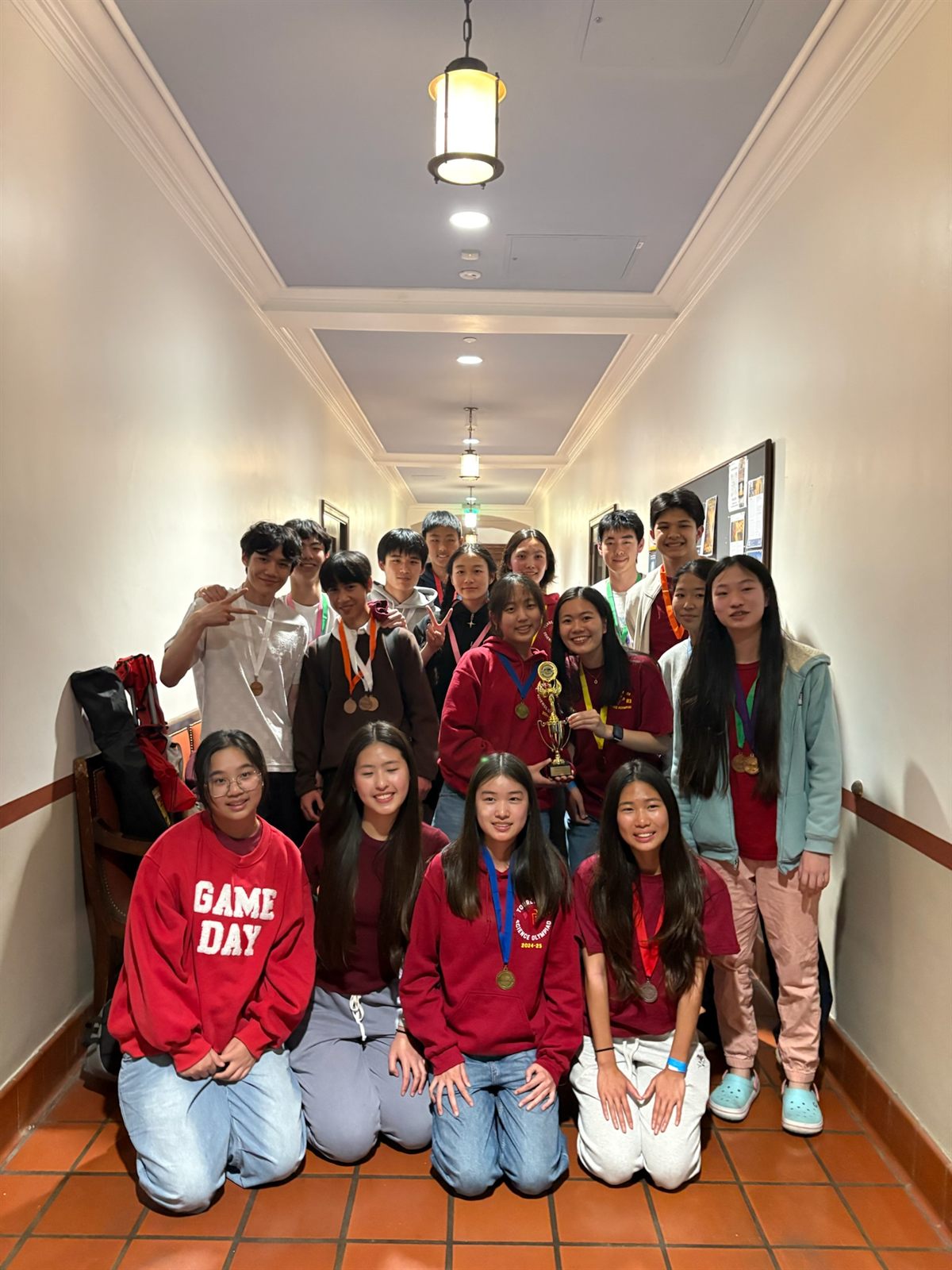Sixteen team members crowded into a tiled hallway after a tournament, medals around their necks and one student holding a gold trophy