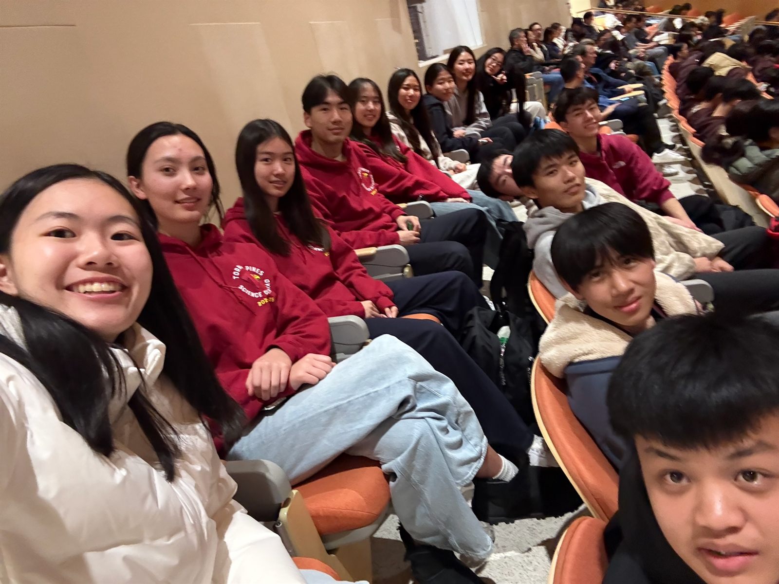 A row of team members in maroon hoodies seated in an auditorium, one leaning into the camera for a selfie