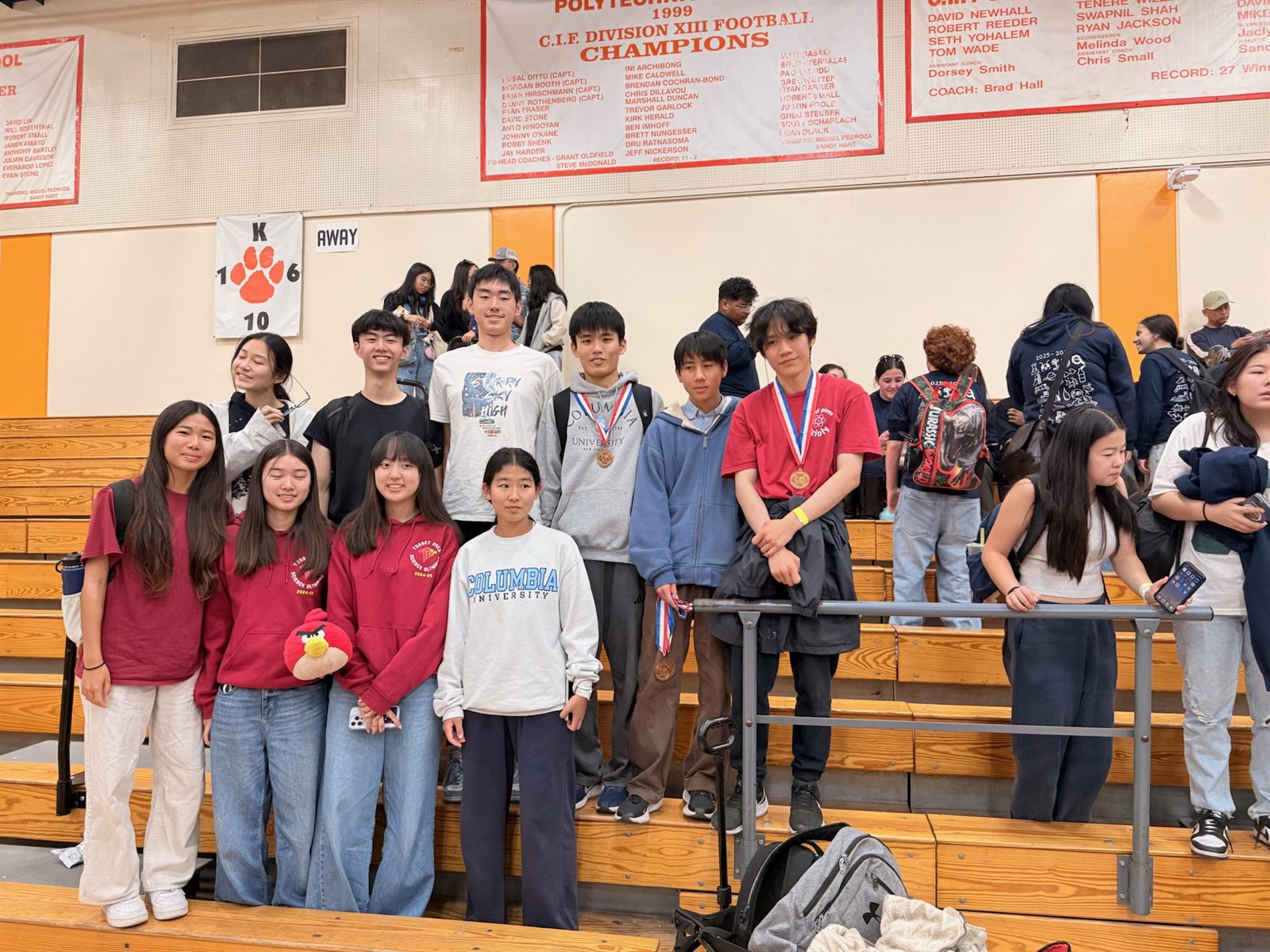 Team members in Science Olympiad hoodies standing on gym bleachers, two wearing medals on red-white-and-blue ribbons