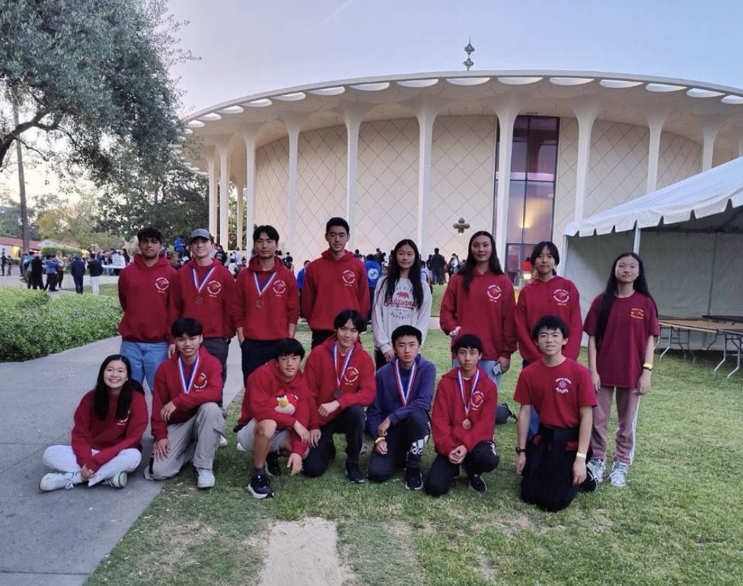 The team in matching red Science Olympiad sweatshirts posed on grass outside a round mid-century building, several wearing medals
